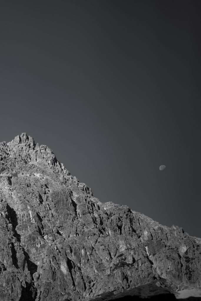 Schwarz-weiß Landschaftsaufnahme von Berggipfeln bei Tageslicht mit sichtbarem Mond am Himmel.
