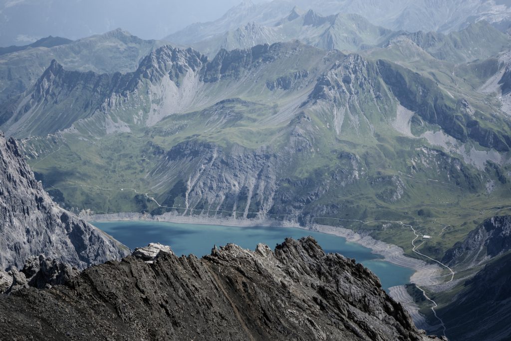 Ein Blick aus der Vogelperspektive auf einen türkisfarbenen Bergsee, umgeben von schroffen, felsigen Gipfeln und grünen Hängen, an deren Hängen kurvenreiche Straßen zu sehen sind.