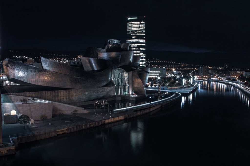 Nachtansicht des beleuchteten Guggenheim-Museums Bilbao mit seiner metallischen, geschwungenen Struktur am Fluss Nervión; im Hintergrund sind die Lichter der Stadt und ein hohes modernes Gebäude zu sehen.