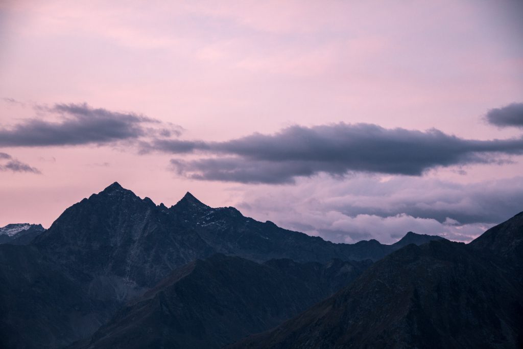 Zerklüftete Berggipfel im Schatten unter einem rosafarbenen und violetten Himmel bei Sonnenuntergang, über dem dunkle Wolken schweben.