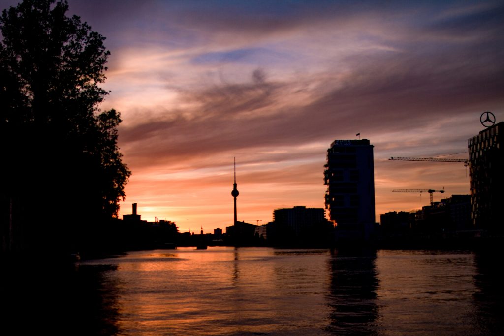 Stadtsilhouette bei Sonnenuntergang mit Silhouetten von Gebäuden, einem hohen Turm, Baukränen und einem Fluss, in dem sich bunte orangefarbene und lila Wolken am Himmel spiegeln.