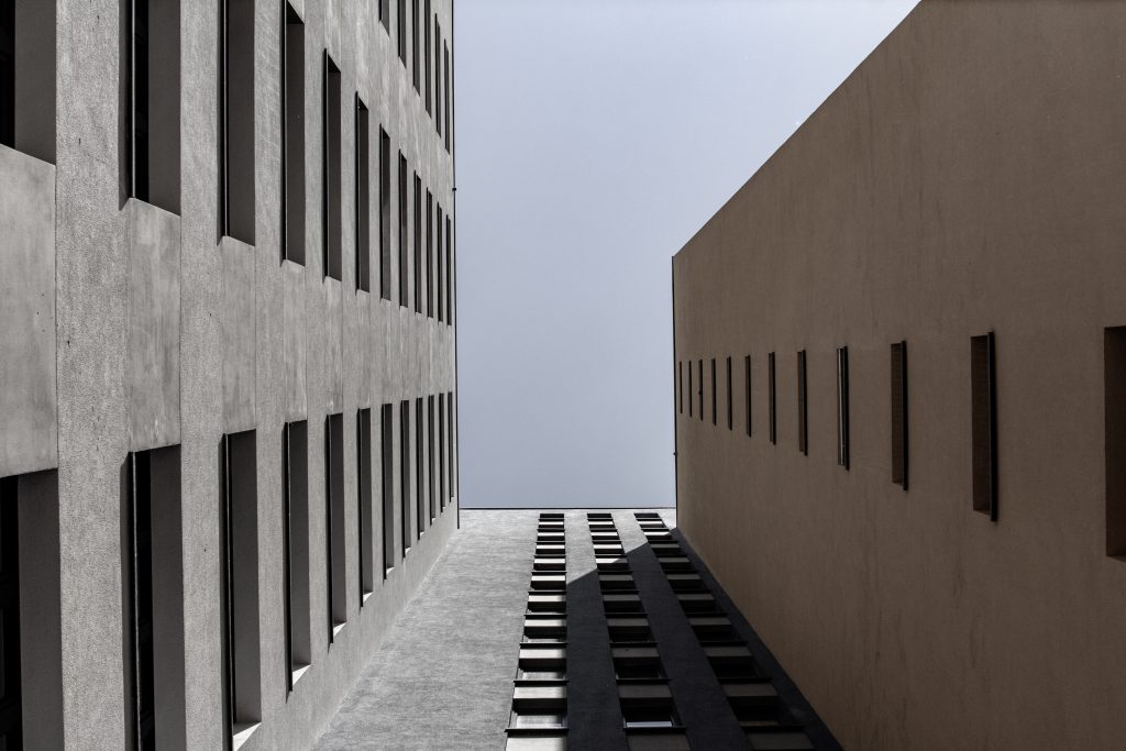 Zwei hohe Gebäude mit Fensterreihen stehen sich gegenüber, so dass eine schmale Gasse zwischen ihnen entsteht, die einen Streifen hellen Himmels über ihnen einrahmt. Schatten und Sonnenlicht heben die geometrischen Linien und Muster hervor.