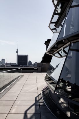 Moderne Glasarchitektur auf der rechten Seite, mit einer Stadtlandschaft mit dem Berliner Fernsehturm und anderen Gebäuden im Hintergrund unter klarem Himmel. Ein Fußweg führt entlang des Bauwerks.