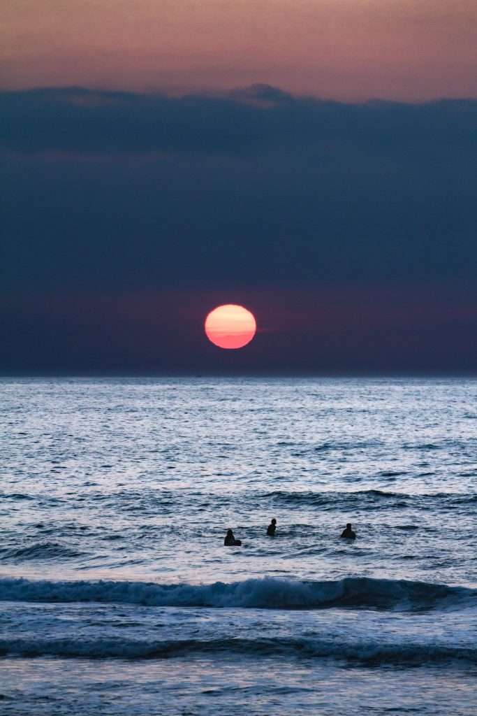 Eine rot-orangefarbene Sonne geht über dem Meer unter und wirft Reflexionen auf das Wasser. Drei Surfer sind in den Wellen zu sehen, die sich gegen den heiteren Abendhimmel abheben.