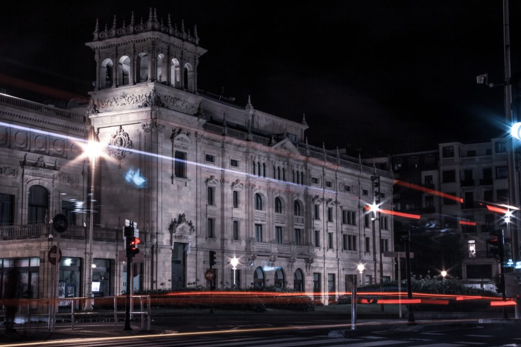 Ein historisches Steingebäude mit verschnörkelter Architektur ist nachts beleuchtet, wobei die Lichtspuren der vorbeifahrenden Autos und Straßenlaternen rote und weiße Streifen über die dunkle Stadtlandschaft ziehen.