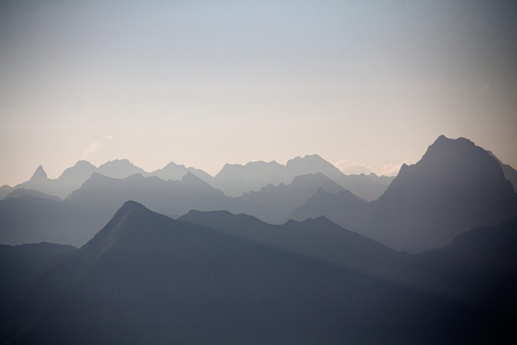 Silhouettierte Gebirgszüge überlagern sich im weichen Licht und bilden Schichten von Gipfeln unter einem dunstigen Himmel. Die Szene wirkt ruhig und beschaulich, mit gedämpften Blau- und Grautönen.