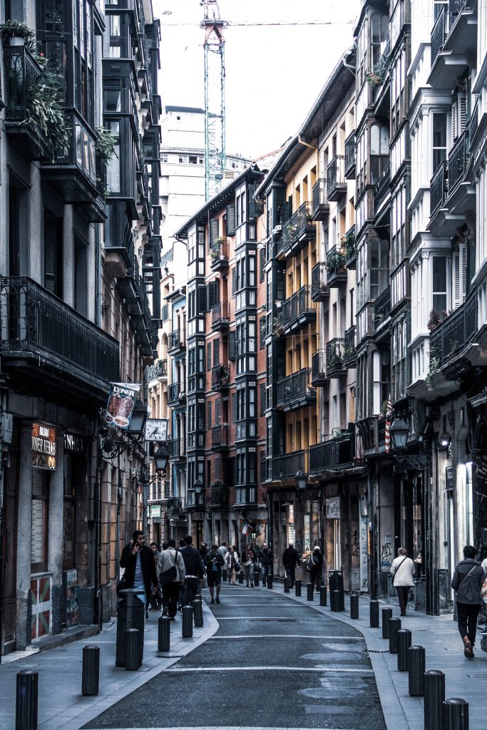 Eine schmale, gewundene Straße mit hohen, bunten Gebäuden mit Balkonen. Die Menschen gehen auf dem Bürgersteig, und Poller trennen den Fußgängerweg von der Straße. Die Atmosphäre ist lebendig und urban.