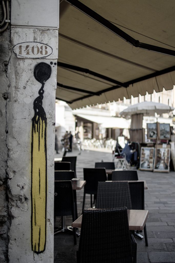 Eine gelbe Kerze mit schwarzer Flamme ist auf eine abgenutzte Säule mit der Aufschrift 1408 gesprüht, die neben Tischen und Stühlen eines Straßencafés unter großen beigen Markisen in einer städtischen Straße steht.