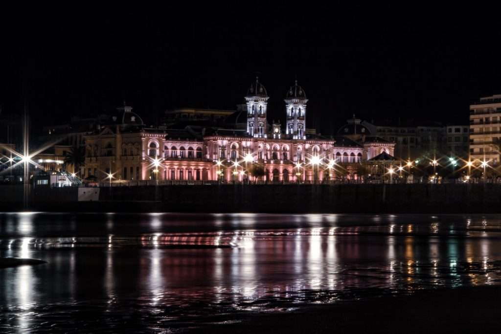 Nächtliche Uferpromenade von San Sebastián mit beleuchteten Gebäuden und reflektierendem Wasser.
