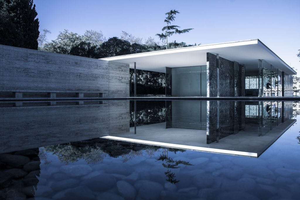 Ein modernistisches Gebäude mit Glaswänden und einem Flachdach spiegelt sich in einem stillen Teich, umgeben von Steinflächen und Bäumen unter einem klaren blauen Himmel.