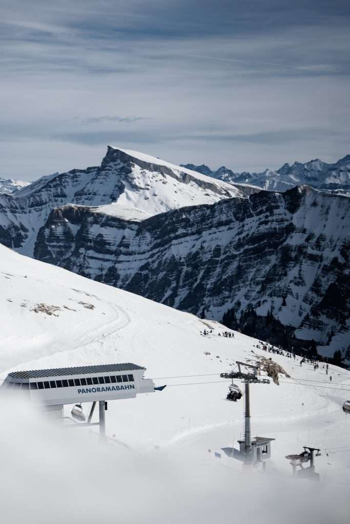 Eine verschneite Berglandschaft mit einem Skilift mit der Aufschrift Panoramabahn, Skifahrern auf der Piste und einem dramatischen, zerklüfteten Berggipfel im Hintergrund unter einem bewölkten Himmel.