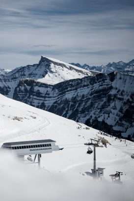 Eine verschneite Berglandschaft mit einem Skilift mit der Aufschrift Panoramabahn, Skifahrern auf der Piste und einem dramatischen, zerklüfteten Berggipfel im Hintergrund unter einem bewölkten Himmel.